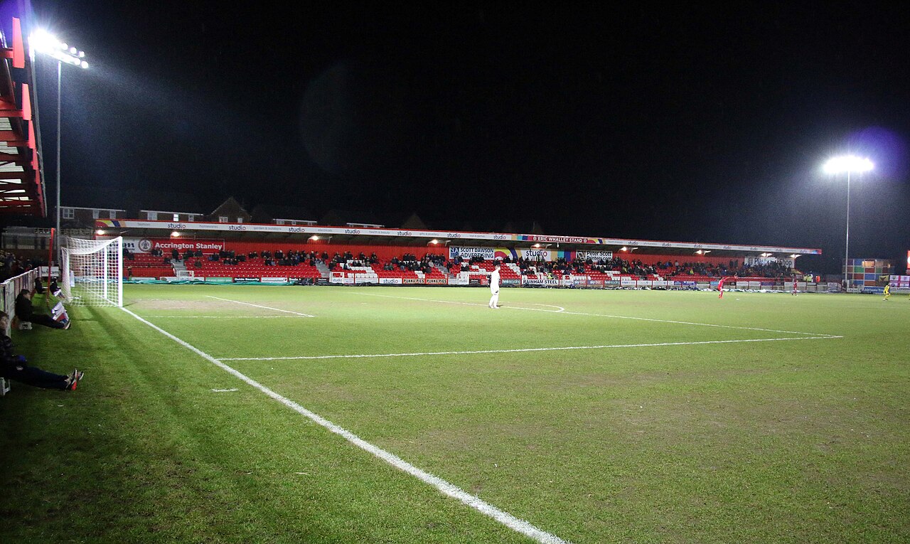 A floodlit evening match at the Crown Ground seen from behind the goal: a single low covered stand of red seats along the far side with a modest crowd in it, advertising hoardings reading Accrington Stanley, two floodlight pylons and dark sky above