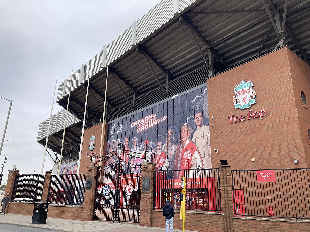 La grada del Kop en Anfield vista desde la calle, con el escudo del Liverpool FC, el Paisley Gateway y la entrada de las visitas al estadio