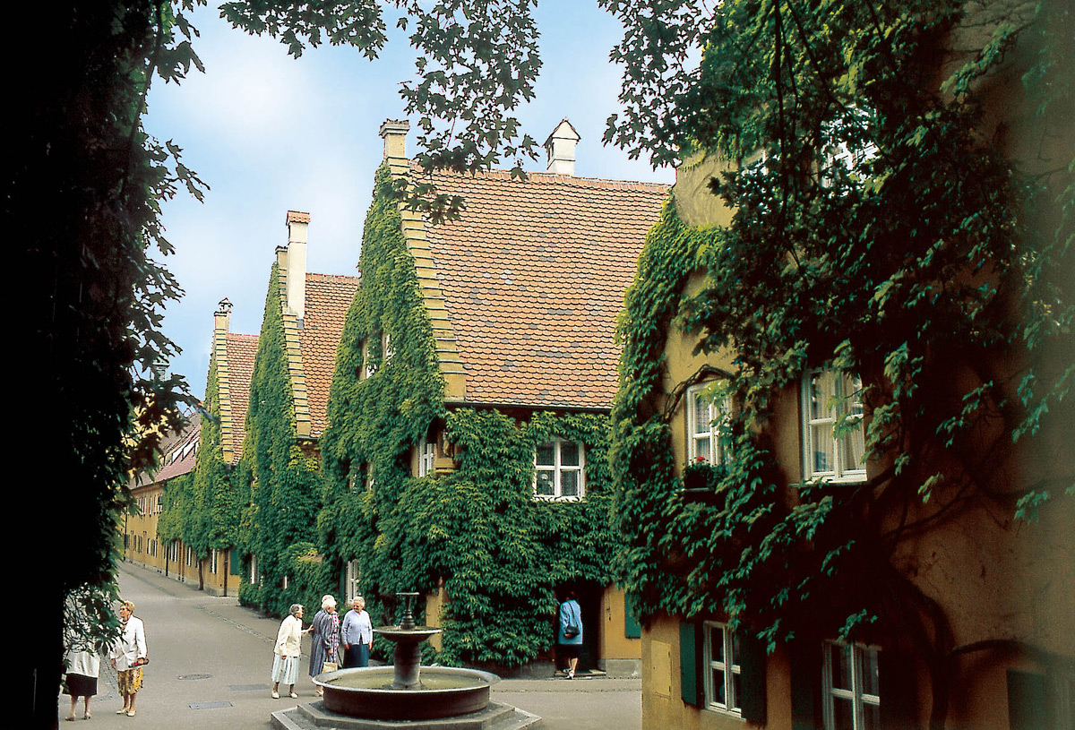 A lane inside the Fuggerei in Augsburg: rows of small yellow houses with steep red tiled roofs and ivy climbing the walls, a stone fountain in the middle of the street and several elderly residents standing beside it