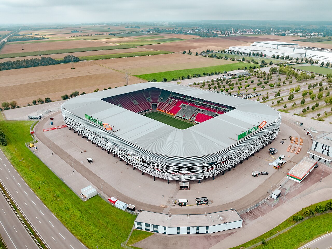 The WWK Arena from the air: a rectangular stadium wrapped in a facade of overlapping white ribbons, with red and pink seats visible inside, standing among ploughed fields with a motorway on one side