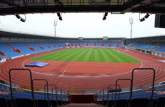 The inside of the Vítkovice municipal stadium seen from a covered stand: blue and red seats, a green pitch in the middle, and a full red running track separating the stands from the touchline