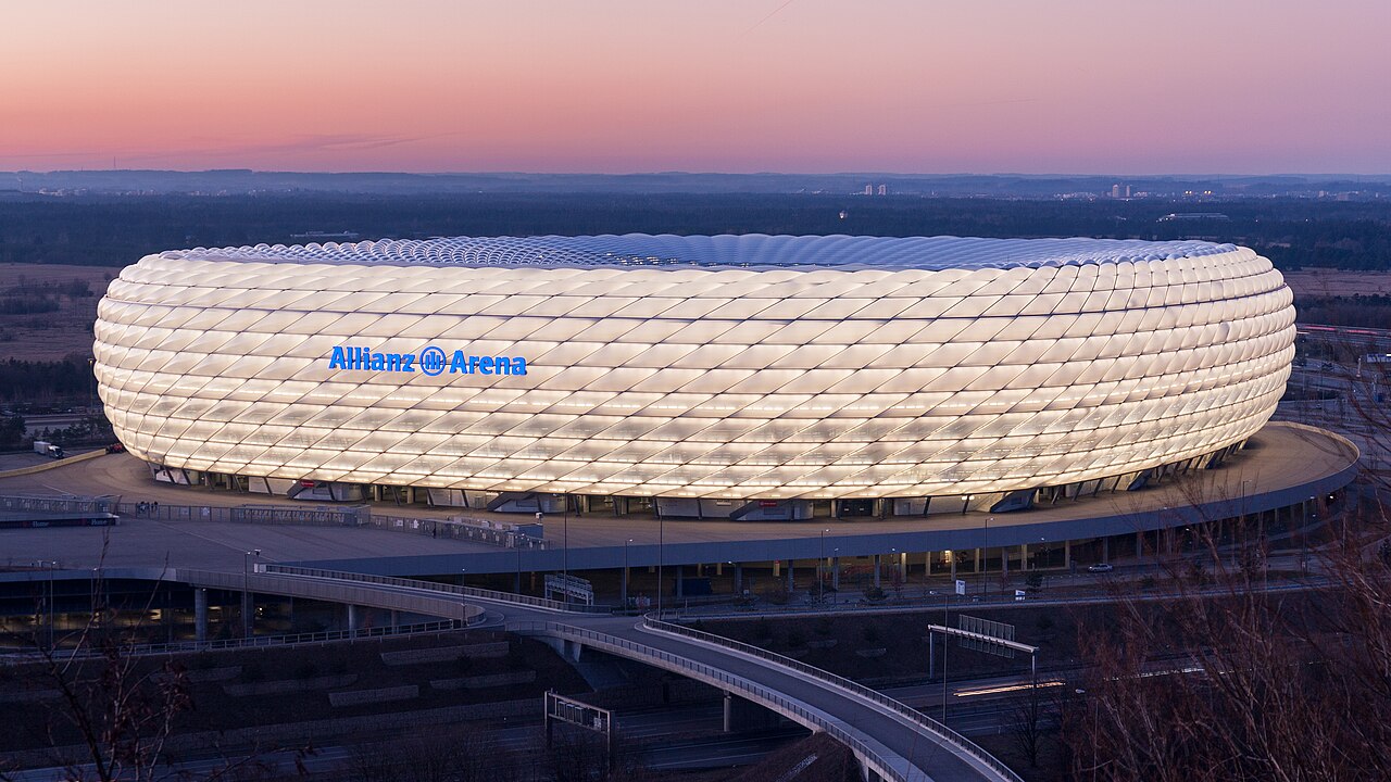 The Allianz Arena at dusk: a huge oval building clad in glowing inflated cushions lit from within, with Allianz Arena in blue lettering on the side and an elevated road curving past below