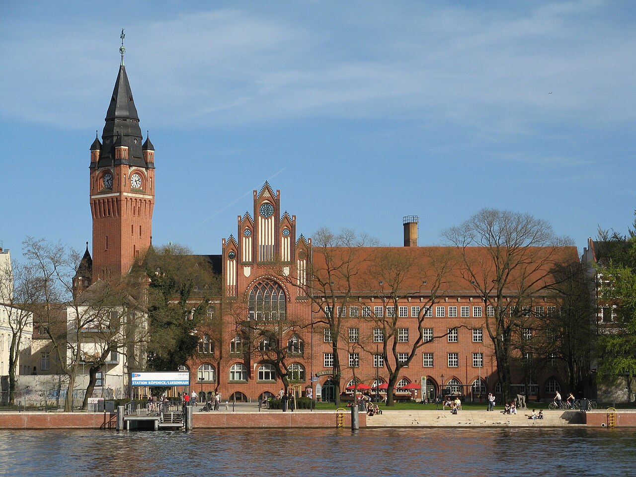 Köpenick town hall seen from across the water: a large red-brick building with a tall clock tower and a steep black spire, bare trees along the bank and people sitting on the steps by the river