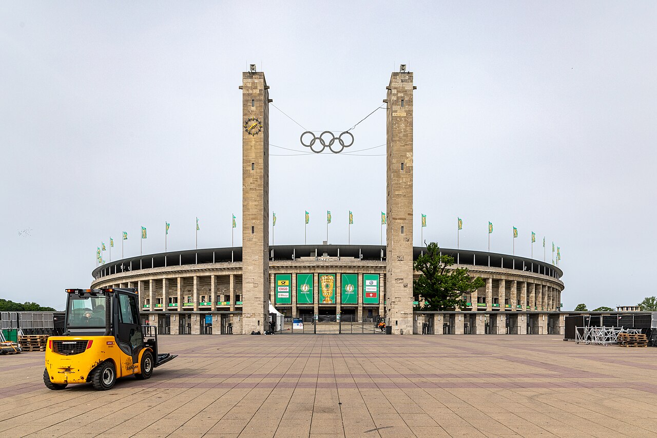 The Olympic Gate at the Berlin Olympiastadion: two tall square stone towers with the Olympic rings suspended between them, the low stone colonnade of the stadium bowl curving away behind, flags along the rim and an empty paved forecourt in front