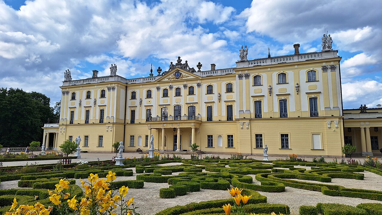 The pale yellow baroque facade of Branicki Palace in Białystok, with statues along the roofline and clipped box hedges and yellow lilies in the formal garden in front