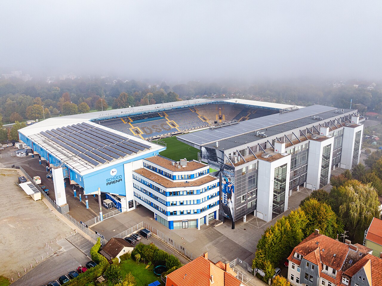 The SchücoArena from the air on a misty morning: a compact stadium with blue and white seats, solar panels covering the roofs of two stands, the Schüco Arena name on the end wall, and ordinary houses with red roofs standing right up against the ground