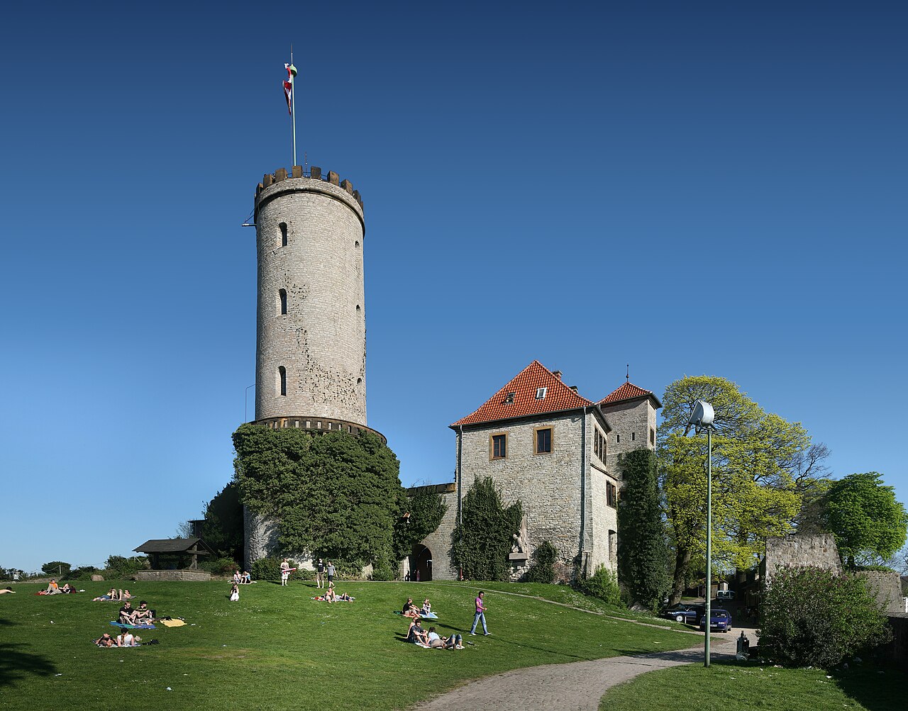 Sparrenburg castle in Bielefeld: a tall round stone keep with battlements and a flag, beside a stone gatehouse with a red tiled roof, with people sitting and lying on the grass slope in front in bright sunshine