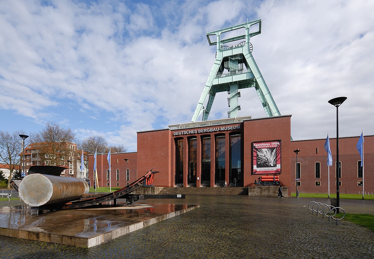The German Mining Museum in Bochum: a low red-brick building with DEUTSCHES BERGBAU-MUSEUM lettering across the front, and behind it a tall pale green steel winding tower with staircases and pulley wheels at the top