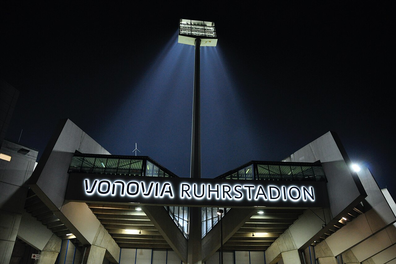 The entrance to the Vonovia Ruhrstadion at night, photographed from below: an illuminated sign reading VONOVIA RUHRSTADION spanning two angular concrete stand structures, with a lit floodlight pylon rising into the dark sky above