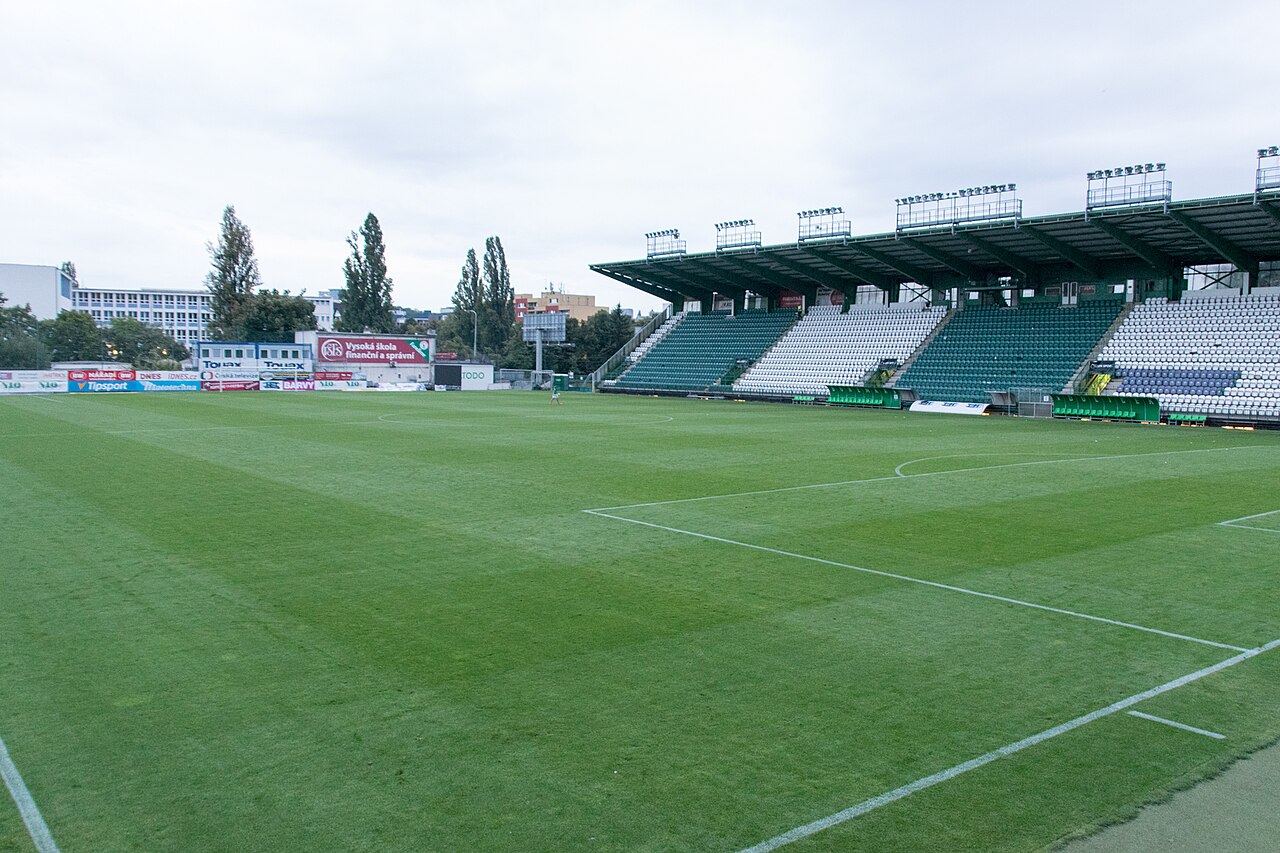 Ďolíček seen from pitch level: a single covered main stand in green and white on one side, low advertising hoardings and trees on the other, and apartment blocks beyond