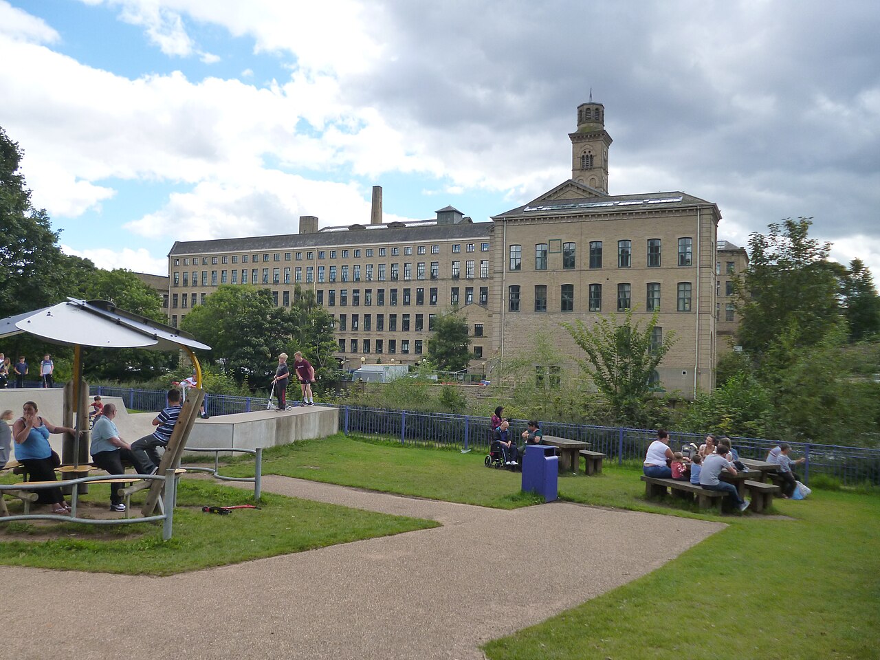 Salts Mill at Saltaire seen across a park: an enormous honey-coloured stone mill of six storeys with rows of tall windows and an Italianate bell tower, with families sitting at picnic tables and children on scooters in the foreground