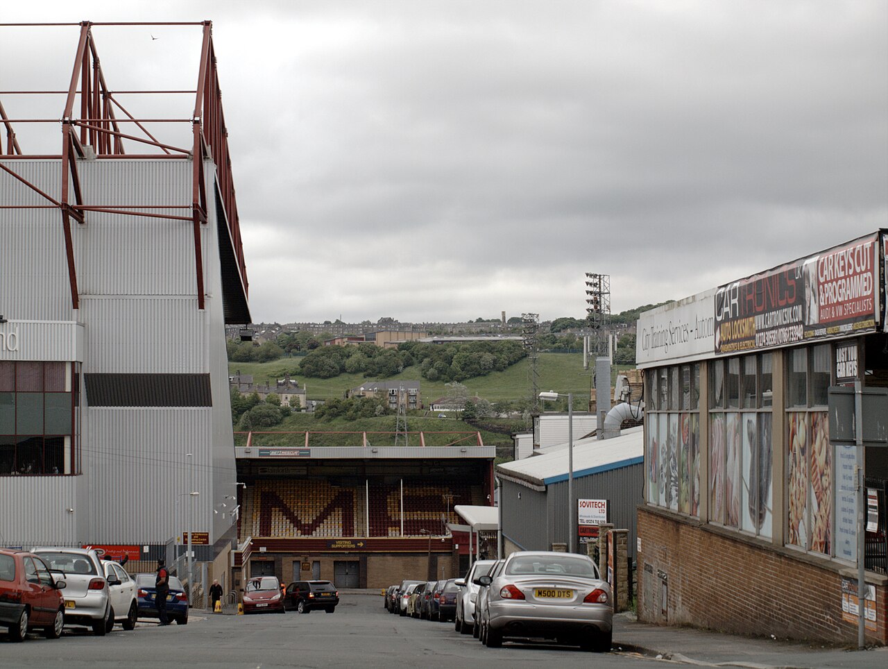 Valley Parade seen along a street: an enormous white main stand with a red steel roof truss rising on the left, the low Bradford End opposite with claret and amber seats and a Visiting Supporters sign, cars parked down the hill and a green hillside of houses rising behind the ground