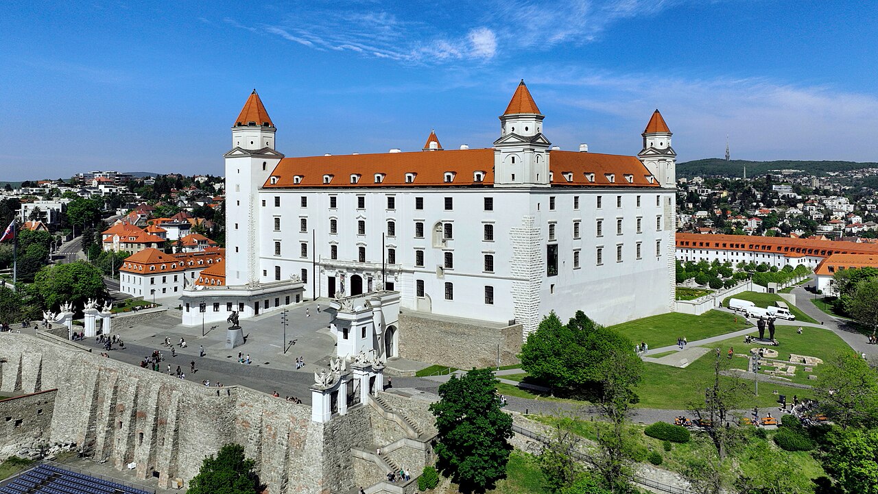 Bratislava Castle from above: a white rectangular palace with red roofs and four corner towers on a hill, with the old town's roofs and the wooded hills of the city behind