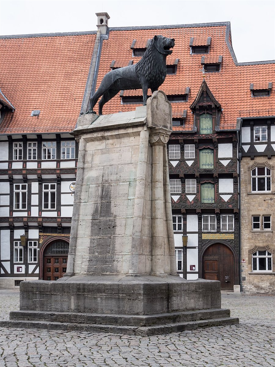 The Brunswick Lion on the Burgplatz: a dark bronze lion with its mouth open standing on a tall pale stone plinth on a cobbled square, with ornately carved half-timbered buildings and steep red-tiled roofs behind it