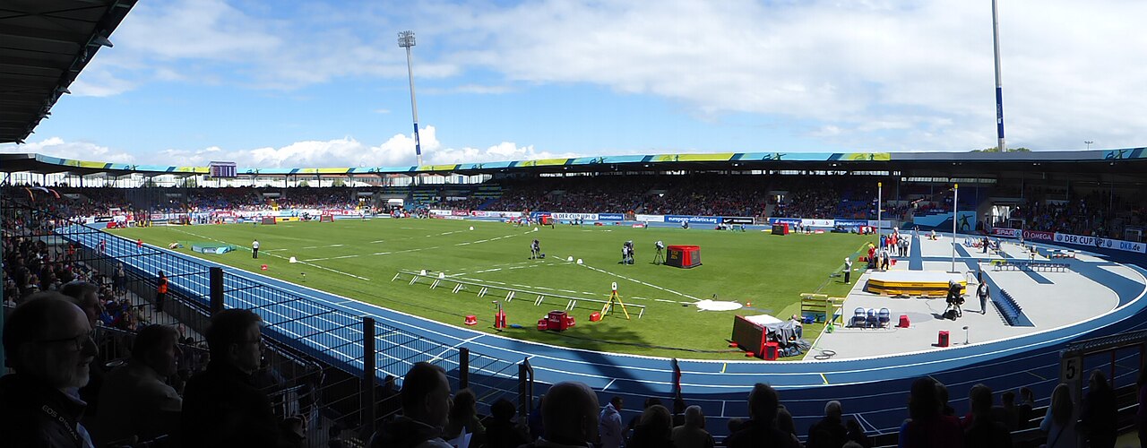 A wide panorama inside the Eintracht-Stadion during an athletics meeting: a blue running track encircling the green infield, hurdles and field-event equipment laid out on the grass, and spectators filling the stands around it