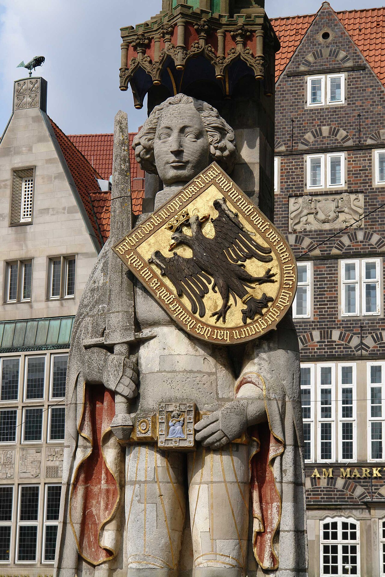 The Roland statue on Bremen's market square in close-up: a stone knight holding a sword and a large gilded shield bearing a black imperial eagle, with gabled merchant houses behind him