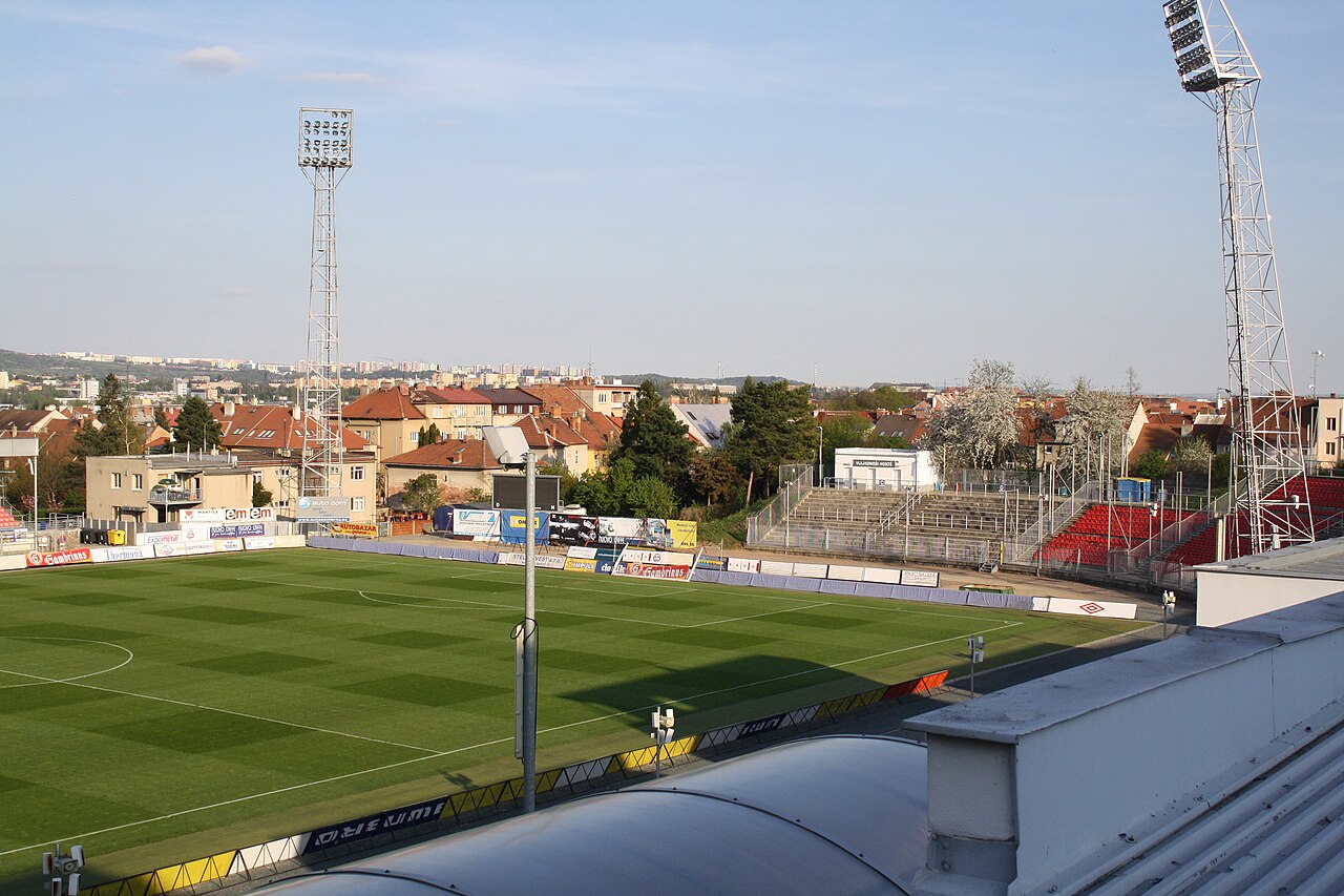 The Srbská stadium in Brno seen from a corner: modest stands with red seats on one side, an open concrete terrace at the far end, tall lattice floodlight pylons and the roofs of Brno behind