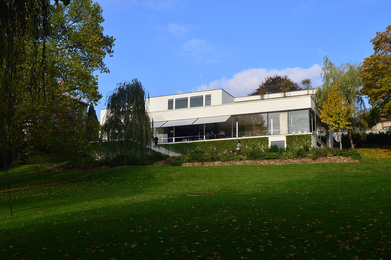 Villa Tugendhat in Brno seen from the lawn below: a white flat-roofed modernist house with a long glazed living room opening onto a terrace, willows and autumn trees to either side
