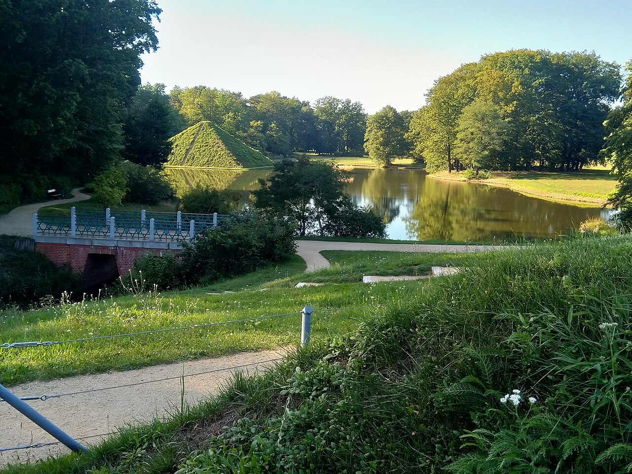 The lake pyramid in Branitz Park near Cottbus: a grass-covered earth pyramid standing at the edge of a still lake and reflected in it, with mature parkland trees around and a path and blue-railed bridge in the foreground