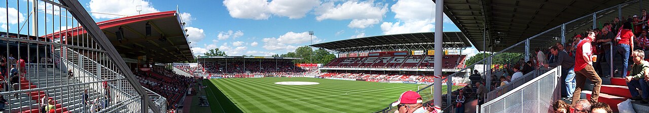 A wide panorama inside the Cottbus stadium: low open stands with red seats on all four sides, the word ENERGIE spelled out across the far stand, open corners, supporters in red scattered through the terraces and a summer sky above