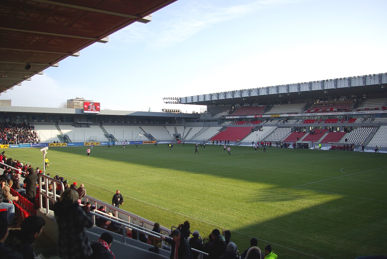 The inside of Cracovia's stadium during a winter match, seen from a corner: red and white seats in four covered stands, players on a green pitch and patches of snow along the touchline