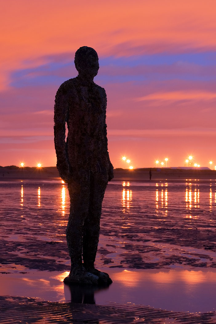 One of Antony Gormley's cast-iron figures on Crosby beach at sunset: a life-size rusted human form standing alone in wet sand, silhouetted against a pink and orange sky, with promenade lights reflected in the shallow water behind and a second figure visible far off