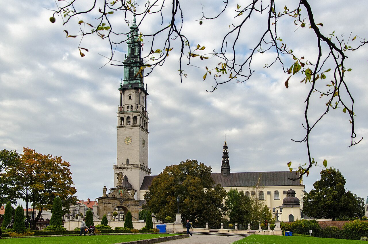 The Jasna Góra monastery in Częstochowa seen from the park: a tall white bell tower with a green copper spire above the basilica and the monastery walls, framed by autumn branches