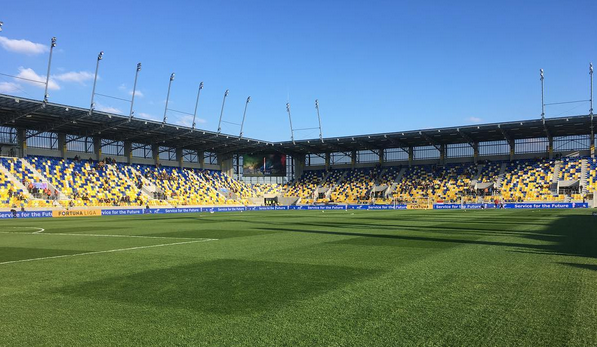 Inside the DAC Aréna: a modern covered stand of yellow and blue seats behind advertising boards reading Fortuna Liga, seen from pitch level across the grass