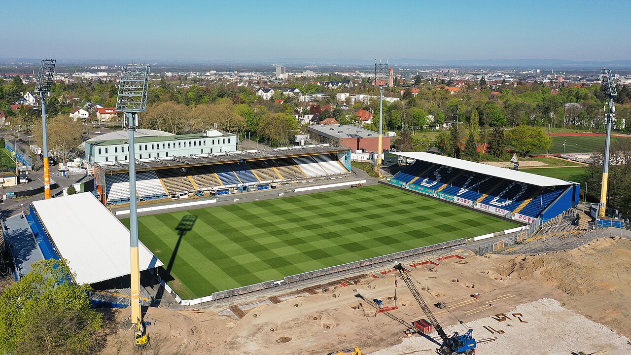 The Böllenfalltor from the air during reconstruction: three stands with blue, white and yellow seats standing around a striped green pitch, one side of the ground reduced to bare earth with a crane and excavated foundations, and the city of Darmstadt spread out behind