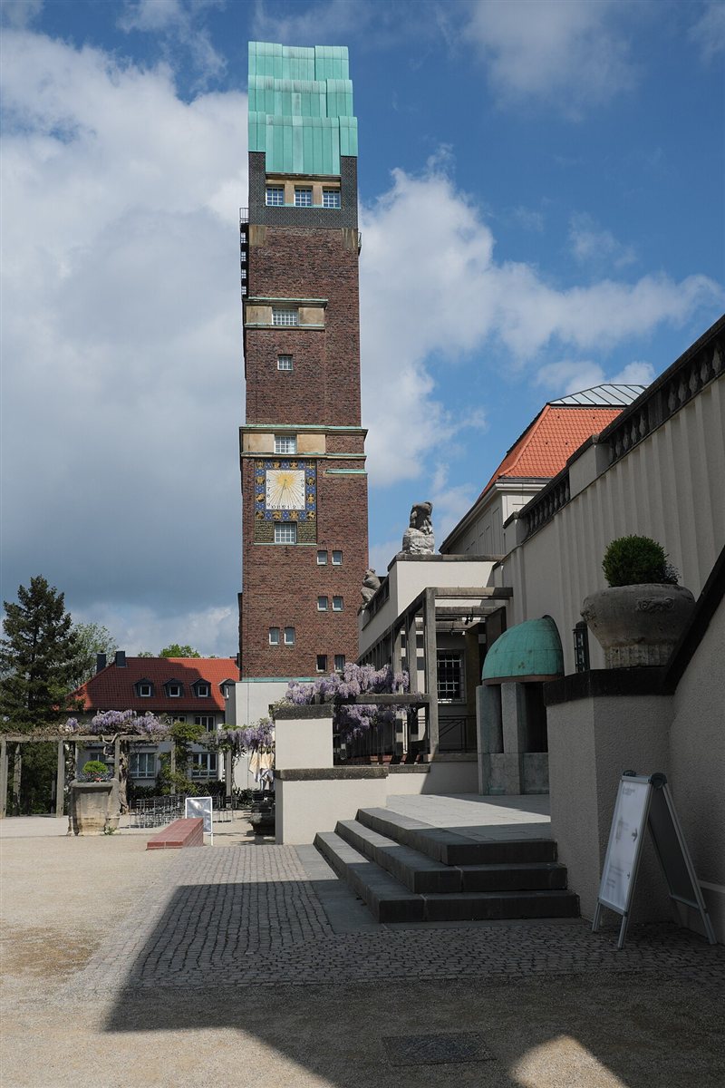The Wedding Tower on the Mathildenhöhe in Darmstadt: a tall brick tower topped by five rounded green copper gables like fingers, with a large mosaic sundial on its face, wisteria in flower on a pergola below and the museum buildings beside it