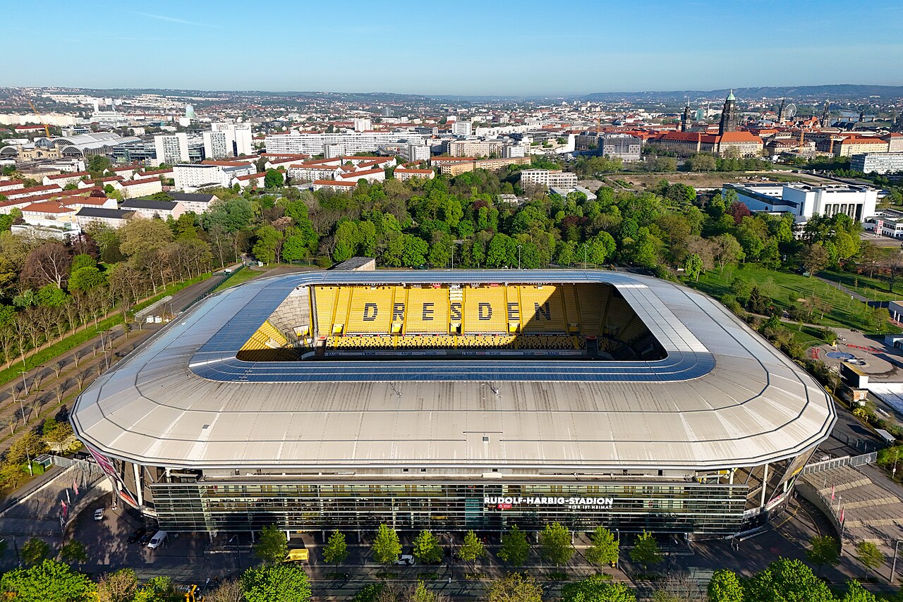 The Rudolf-Harbig-Stadion from the air: a single-tier stadium with a pale grey roof, yellow seats spelling DRESDEN across one stand, parkland around it and the domes and spires of the Dresden old town on the skyline behind