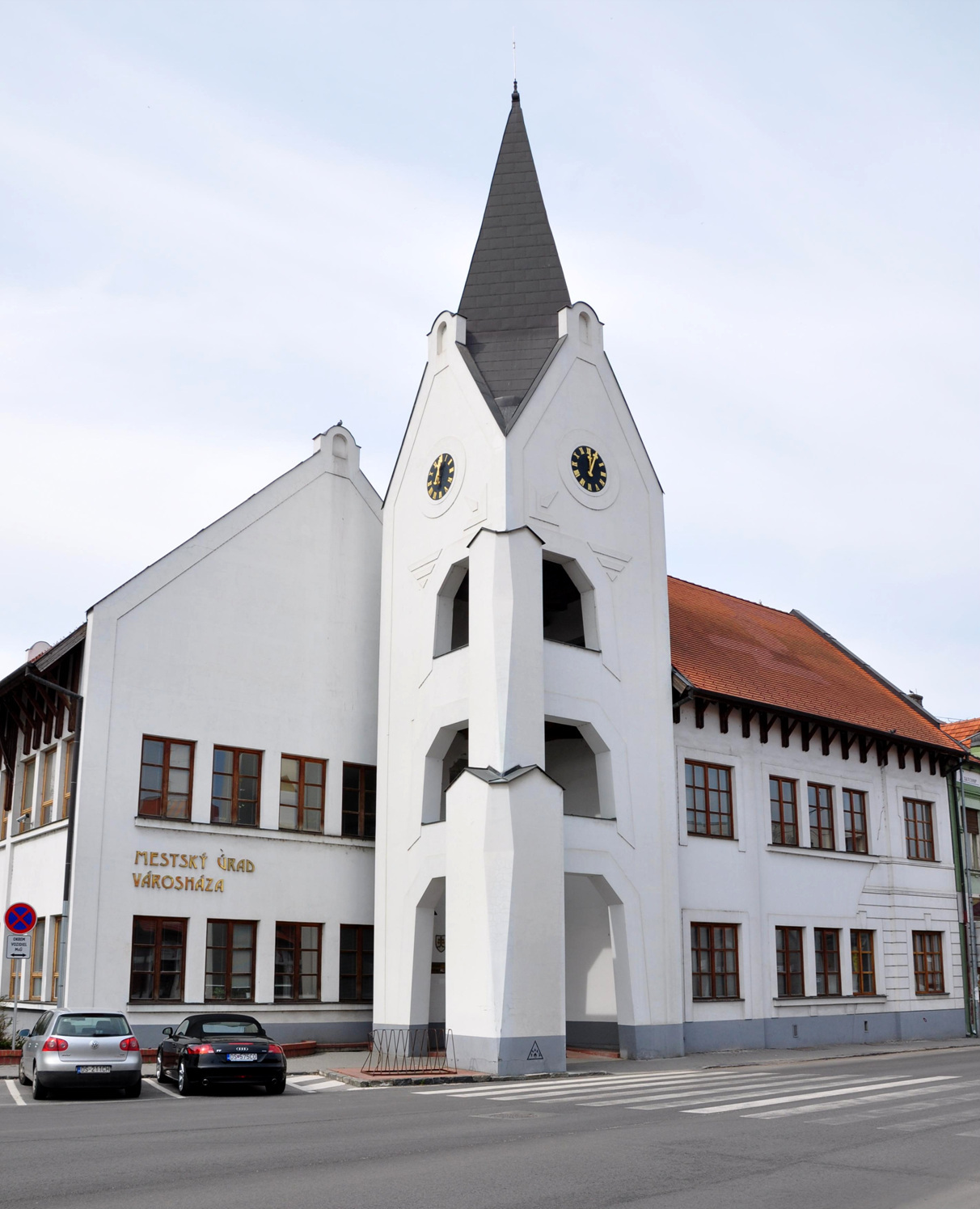 The town hall of Dunajská Streda: a white building with a slate spire and two clock faces, with gold lettering on the wall reading MESTSKÝ ÚRAD above VÁROSHÁZA