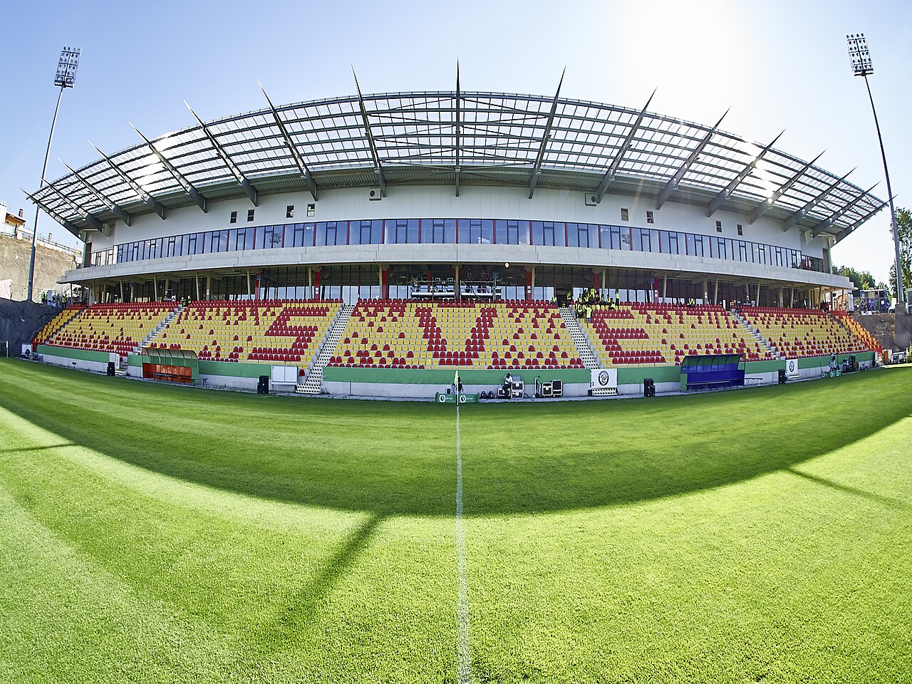 The Ursapharm-Arena an der Kaiserlinde photographed from the pitch with a wide-angle lens: a single new main stand of red and yellow seats under a steel and glass roof, floodlight pylons at each corner and bare earth still visible behind the stand where work continues