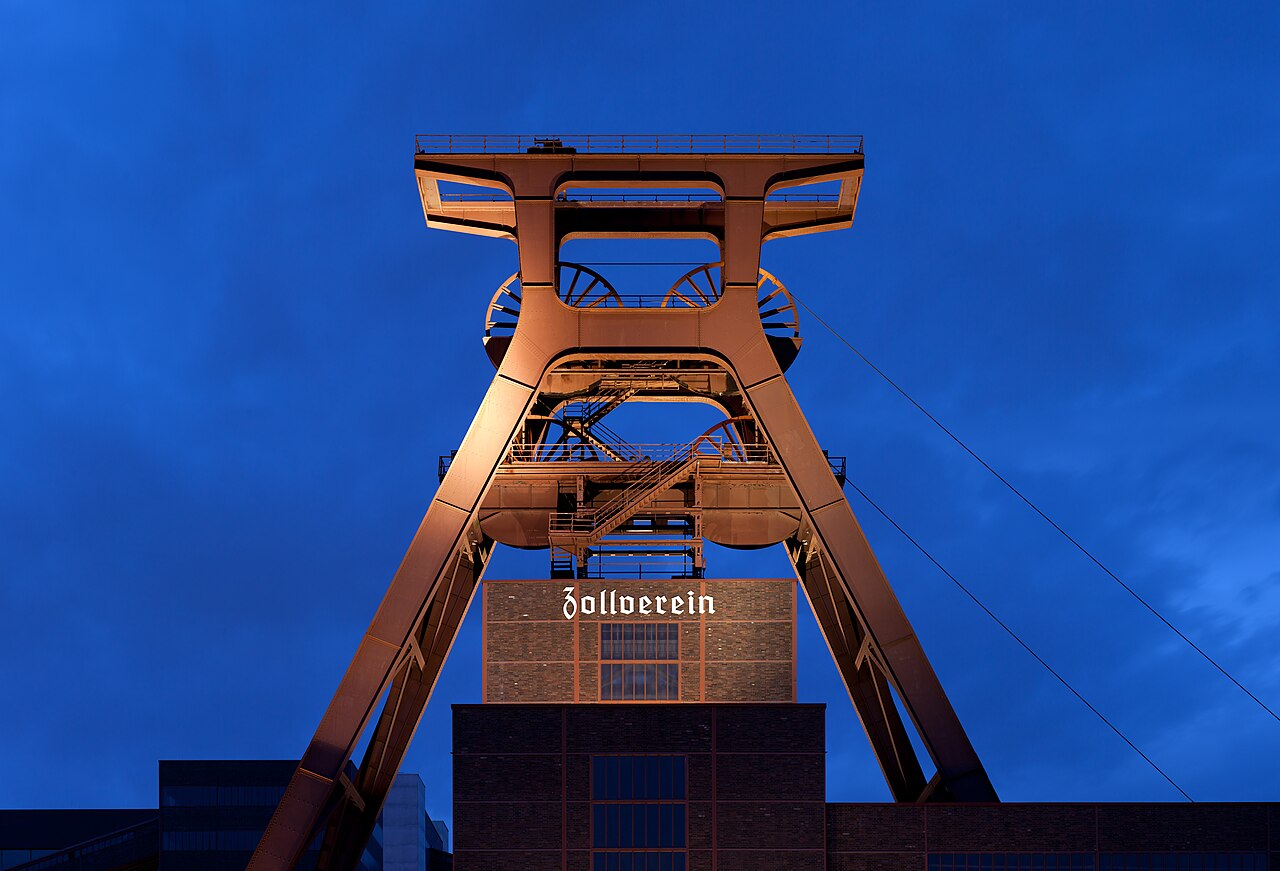 The winding tower of Zeche Zollverein in Essen lit red against a deep blue evening sky, with the word Zollverein in Gothic lettering on the brick building beneath it