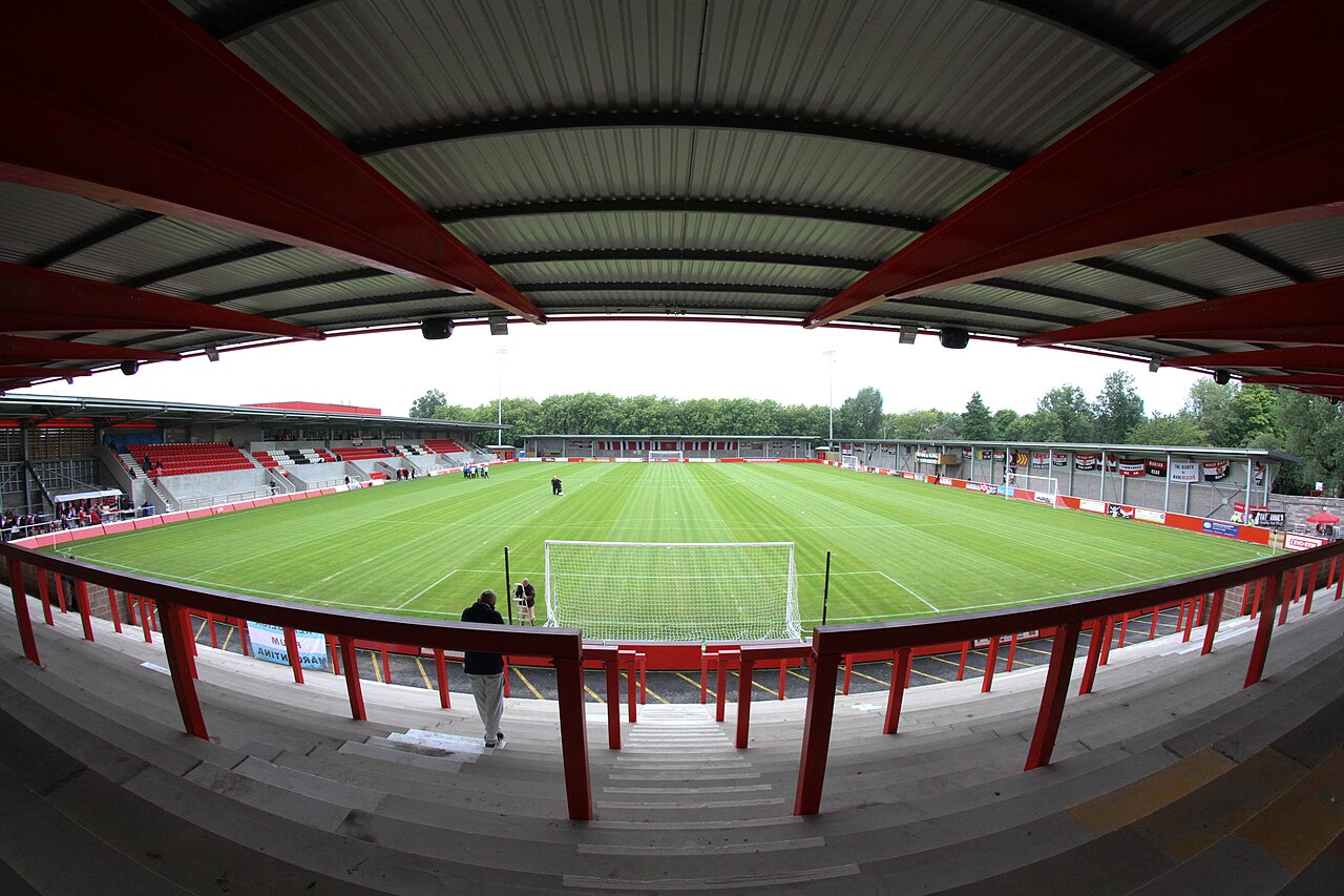 Broadhurst Park seen from the terrace behind a goal through a wide-angle lens: red painted crash barriers and steelwork in the foreground under a corrugated roof, a low four-sided ground with red and white seats and terracing all round, and trees behind the far stand