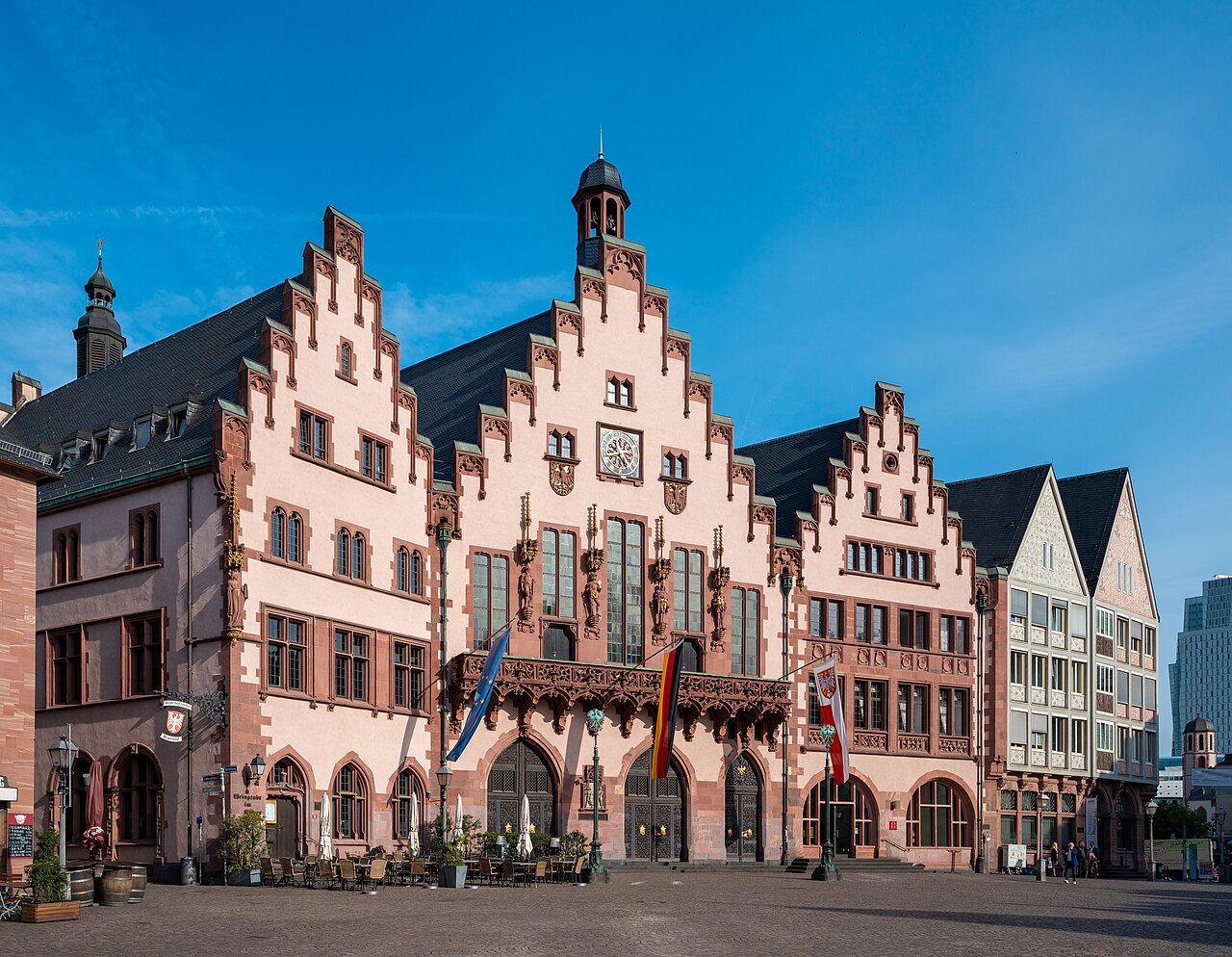 The Römer in Frankfurt: three pink-and-red stepped-gable town hall houses with a small bell turret, flags hanging over the arcade, café tables on the cobbles and a modern glass tower visible at the right edge