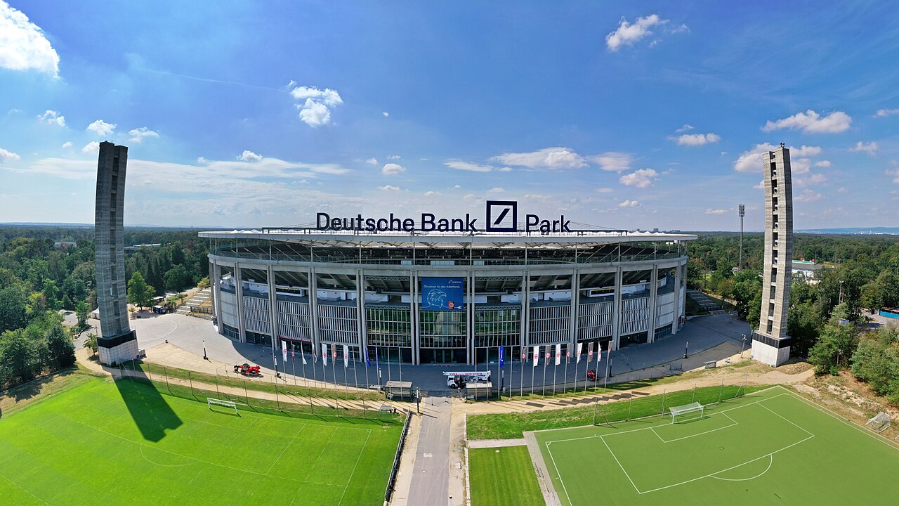 The Waldstadion in Frankfurt seen from the air: a round stadium with Deutsche Bank Park lettering on the roof, two tall concrete floodlight towers flanking it, and forest stretching away on all sides