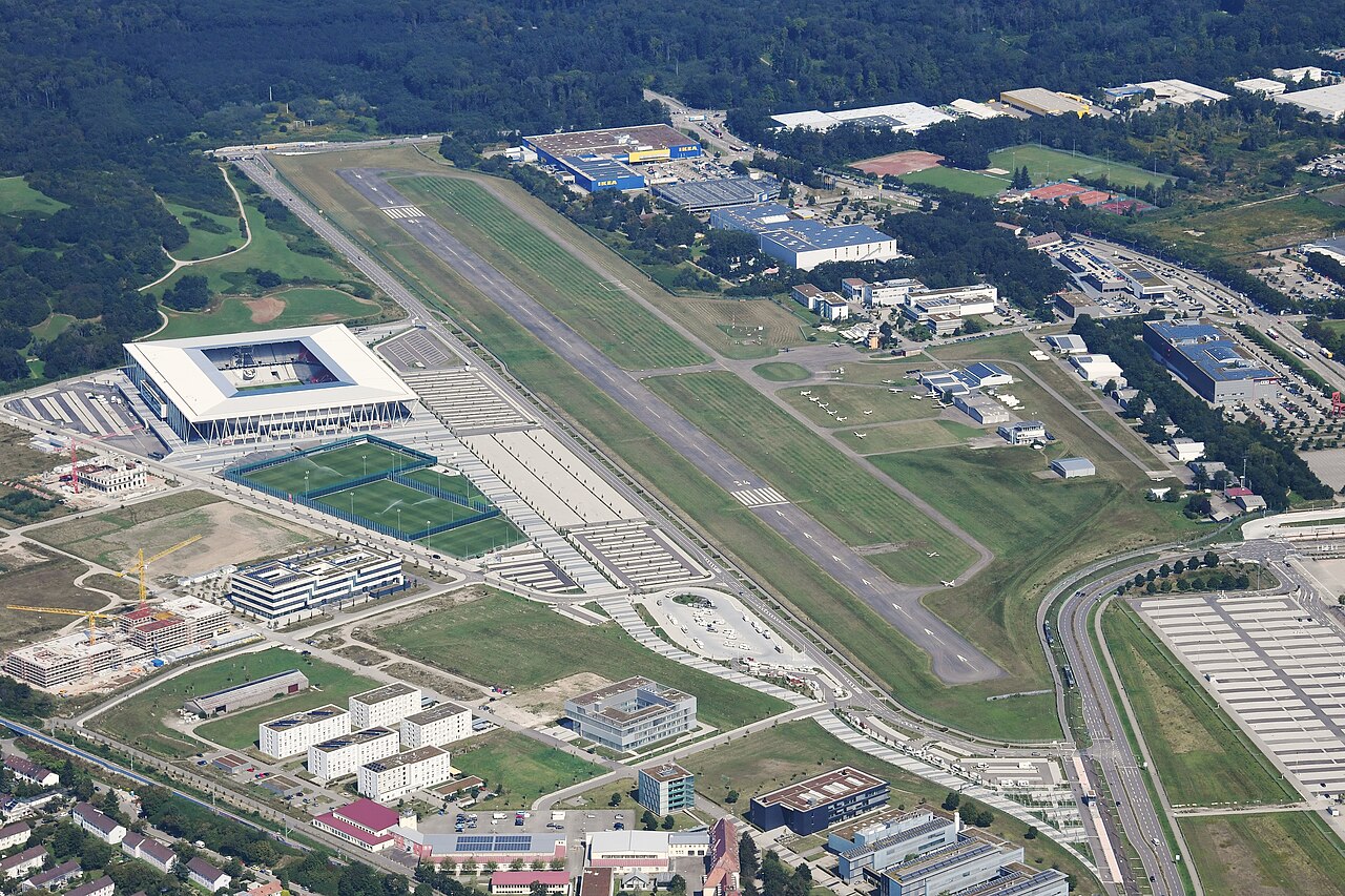 Aerial view of the Europa-Park Stadion standing directly beside Freiburg's airfield: the low white stadium on the left, the runway and parked light aircraft on the right, with training pitches between them