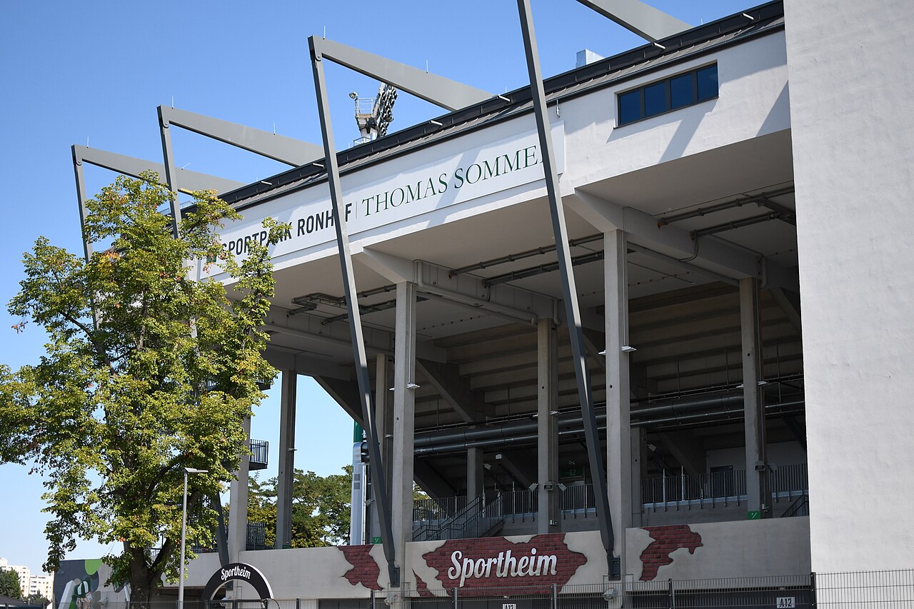 The main stand at the Sportpark Ronhof: a concrete stand with angled steel roof supports and the lettering SPORTPARK RONHOF THOMAS SOMMER along its front edge, a floodlight pylon behind, and a Sportheim sign at ground level
