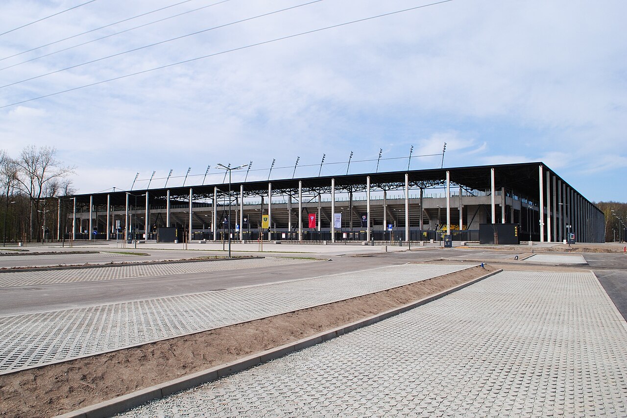 The Arena Katowice from outside shortly after opening: a long single-tier stand under a flat black roof on slender columns, with a newly laid car park of grass pavers in front and woodland behind