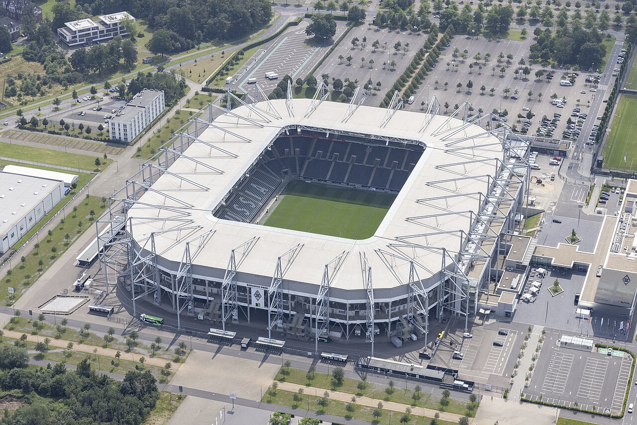 Borussia-Park from the air: a modern enclosed stadium with a white roof carried on a ring of steel struts, surrounded by very large tree-lined car parks on the edge of town