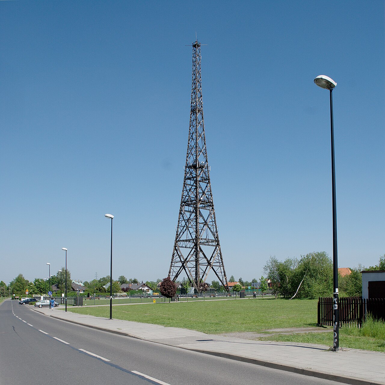 The Gliwice radio tower: a tall dark wooden lattice mast tapering to a point, standing alone on a grass field beside an ordinary suburban road under a clear blue sky