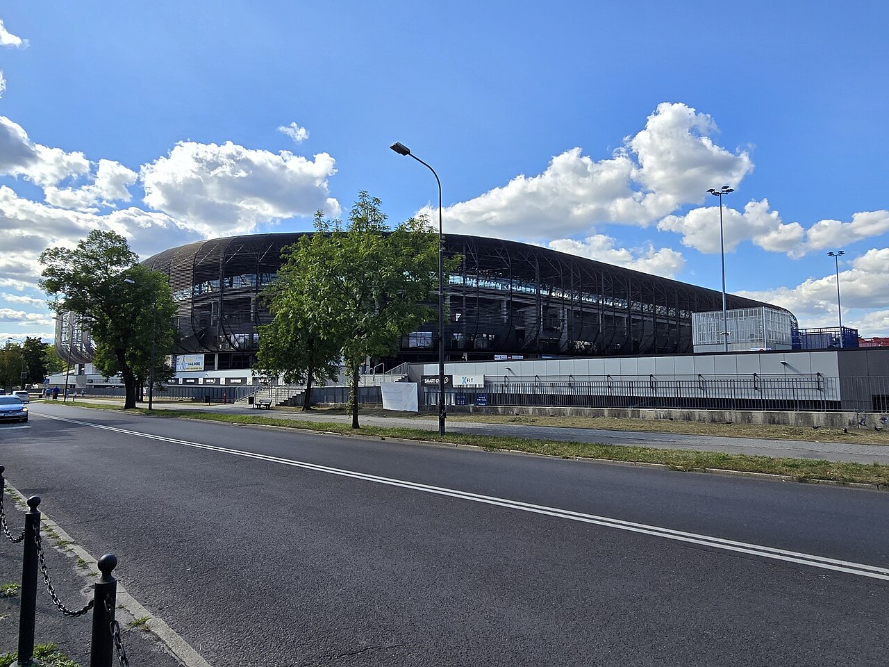 Arena Zabrze seen from Roosevelta Street on a sunny day: a dark, curved stand with an open lattice roof structure rising behind trees and a low perimeter wall