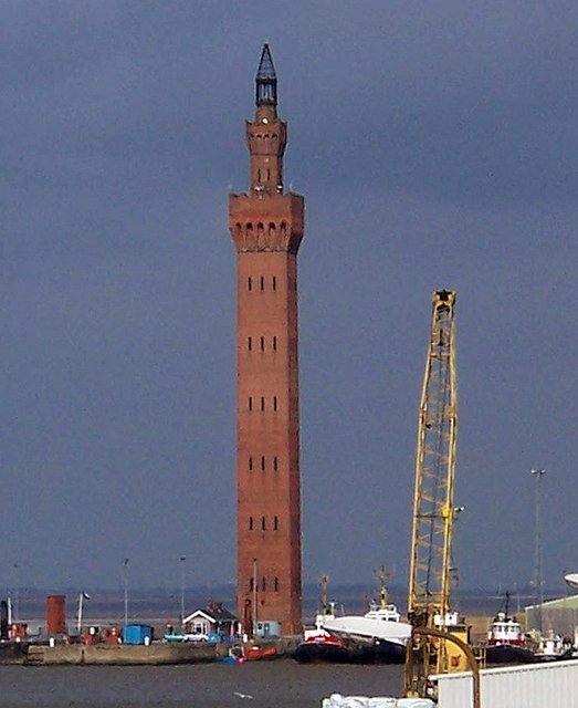 Grimsby Dock Tower against a dark storm sky: a tall slender red-brick Italianate tower with an arcaded top stage and a small spire, rising above the fish docks with a yellow crane, fishing boats and low dock buildings below it
