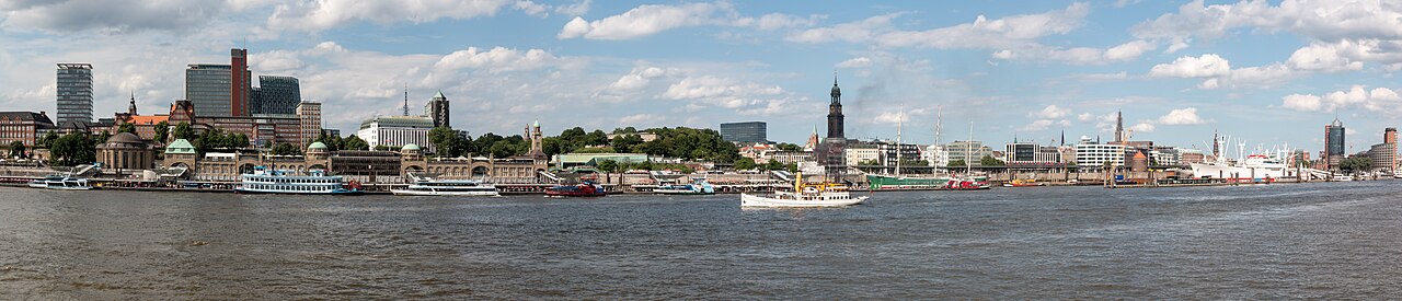 The Hamburg waterfront seen in a wide panorama from across the Elbe: the long Landungsbrücken terminal with its copper domes and clock tower, paddle steamers and ferries moored along the piers, the tall spire of the Michel church behind, and harbour cranes stretching away to the right