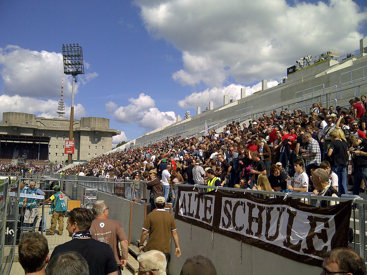 A packed standing terrace at the Millerntor: supporters in brown and white behind a hand-painted banner reading Alte Schule, a floodlight pylon above them, and behind the stand the grey concrete mass of the Heiligengeistfeld flak tower with Hamburg's television tower beyond