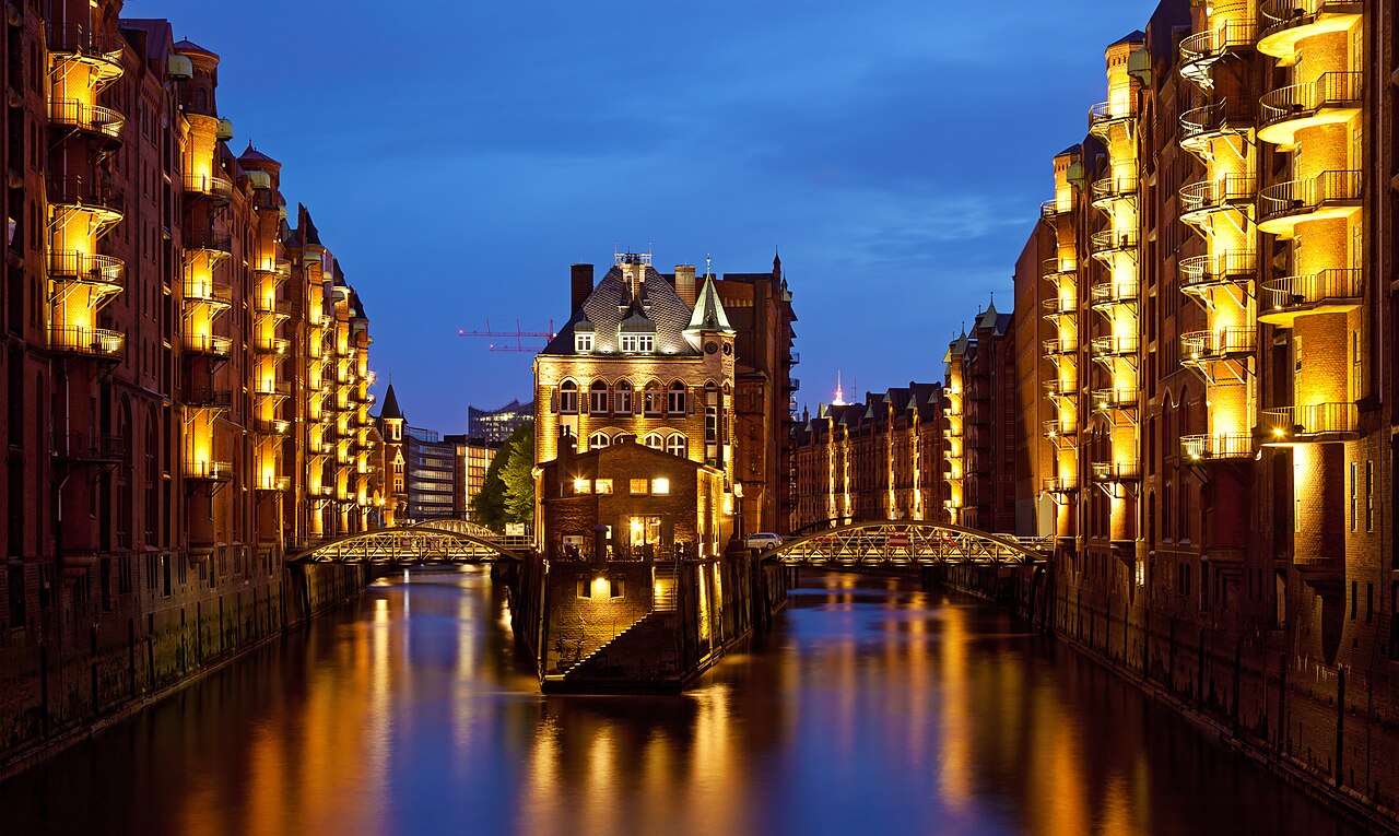 The Speicherstadt in Hamburg at dusk: tall red-brick warehouses lit gold along both sides of a canal, iron footbridges crossing it, and the whole scene reflected in still water