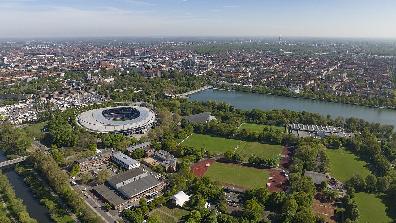 The Niedersachsenstadion from the air: an oval stadium with a pale grey roof standing among trees, the long Maschsee lake beside it, and the city of Hanover with the domed Neues Rathaus spreading out behind