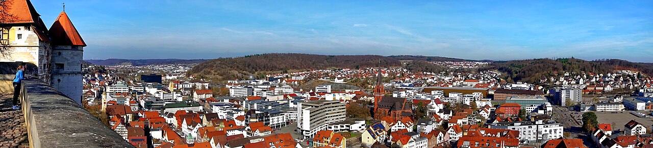A wide panorama of Heidenheim seen from the wall of Schloss Hellenstein: a visitor leaning on the parapet in the foreground, the red roofs of the town spread out below with a church spire in the middle, and wooded hills of the Swabian Alb closing the horizon