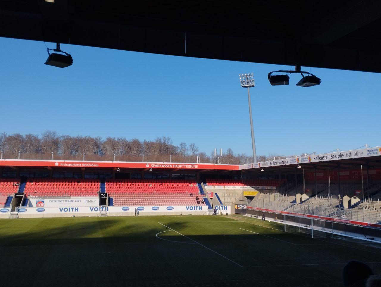 Inside the Voith-Arena seen from under a stand roof: a low main stand of red seats with Sparkassen Haupttribüne lettering along its roof, VOITH advertising on the pitchside hoardings, a floodlight pylon, and a bare wooded slope rising directly behind the far stand
